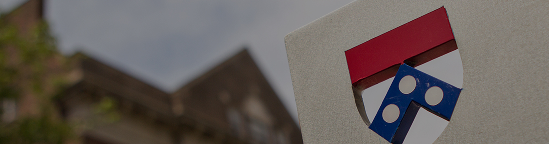 A partial view of a university emblem on a sign with a blurred building and tree in the background.